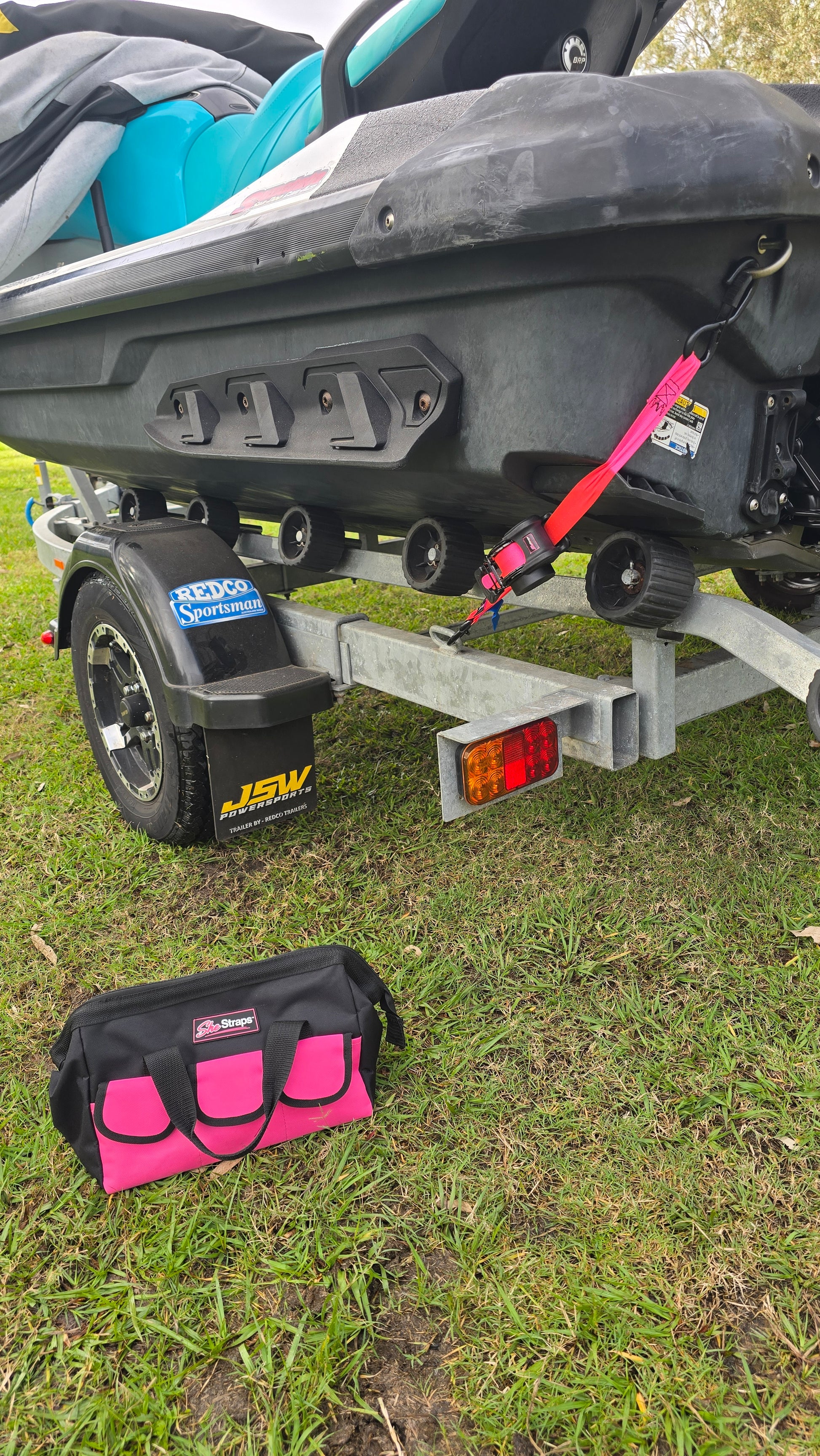A close-up outdoor shot of a black and gray and turquise jetski and trailer assembly with a pink strap, next to a matching pink and black bag on grass.