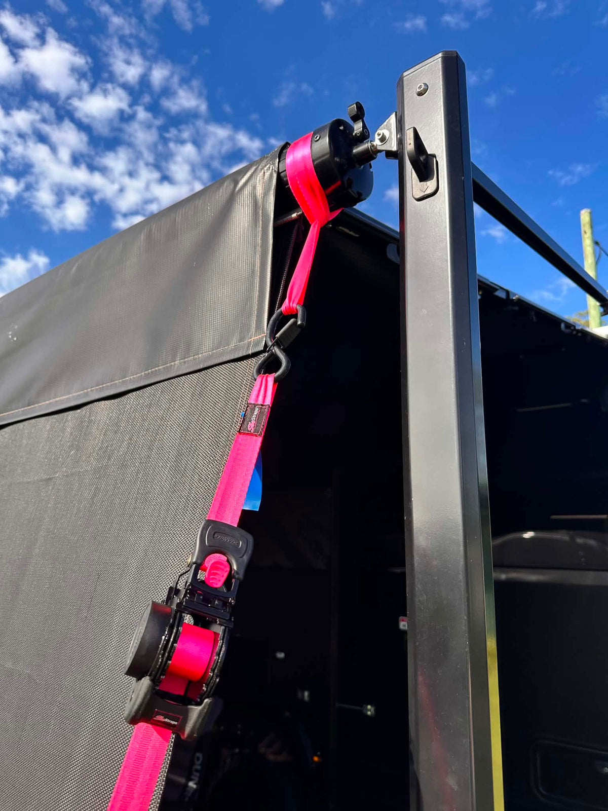 Bright pink and black vehicle window tie-down strap secured to a black metal frame  caravan awning under a blue sky with white clouds.