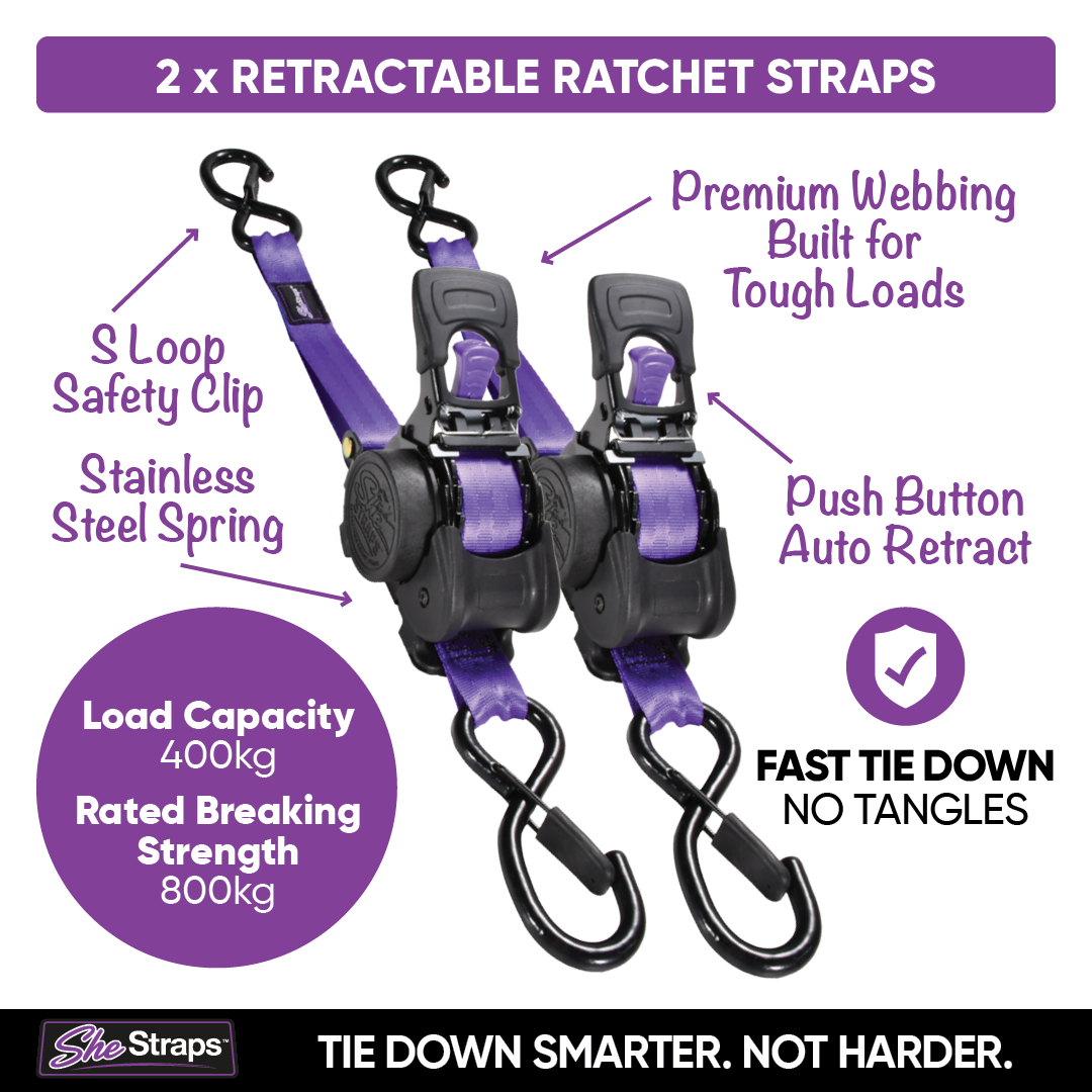 Two purple and black retractable ratchet straps with callout labels on a white background, including text about S-loop safety clip, stainless steel spring, push button auto retract, load capacity, and fast tie down no tangles. SheStraps logo at the bottom.